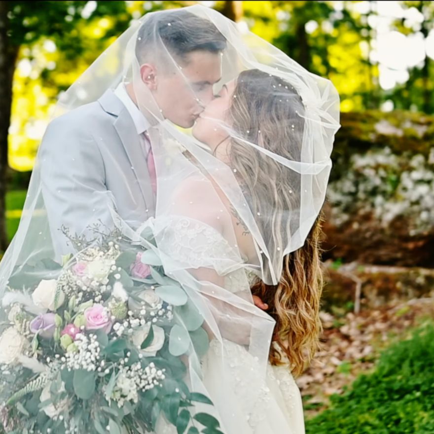 A couple kissing under the bride's veil in a sunlit wood