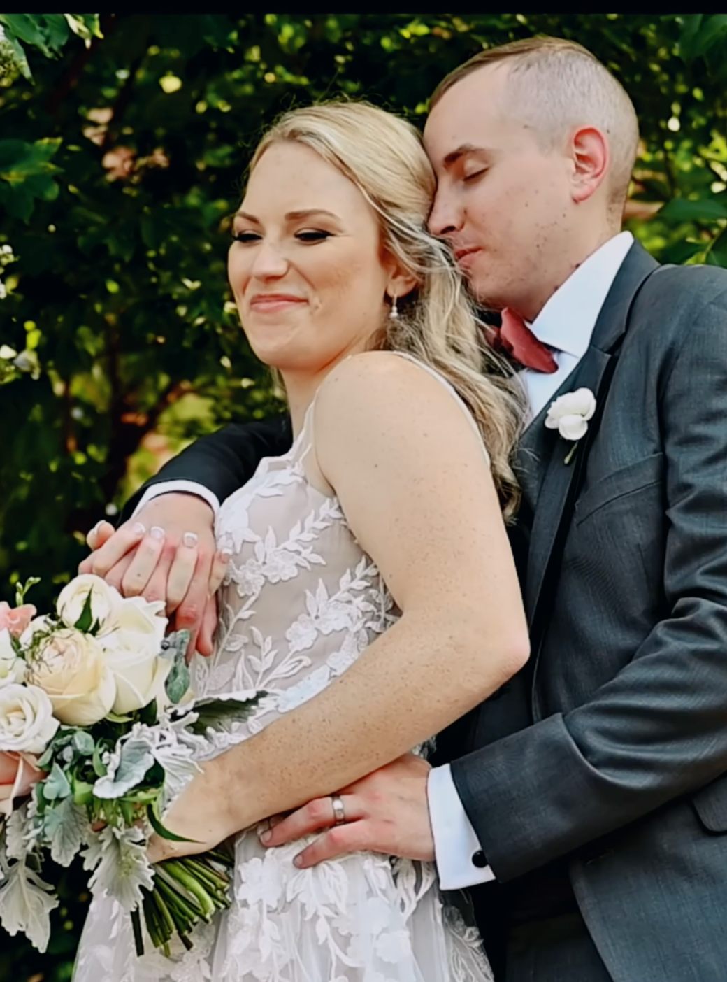 A bride smiling as her groom leans in close, framed by flowering garden branches