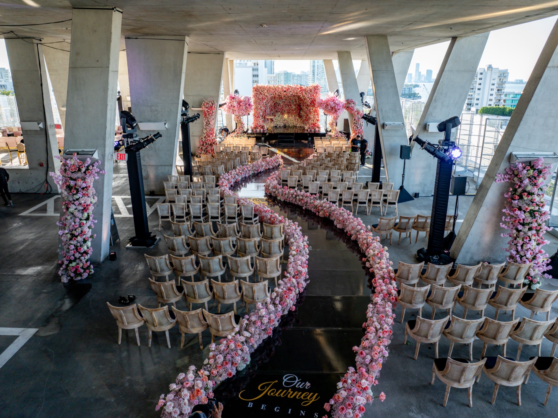 An empty ceremony space seen from above, a curving aisle of blush roses winding between rows of chairs toward a full floral backdrop